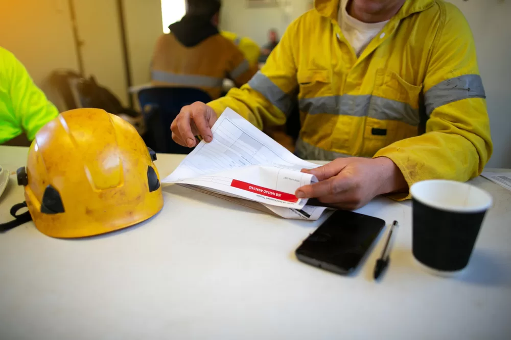 Planner site engineer sitting placing safety hard hat on the table coffee cup pen reviewing JSA risk assessment working at height document prior sign approvals permit by senior site supervisor
