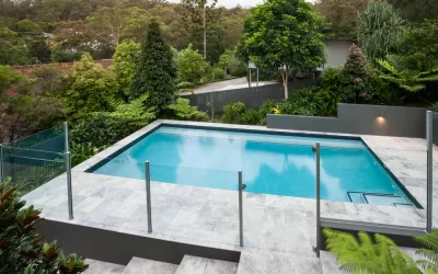 Modern swimming pool with a glass fence on the tile floor covered with a garden with green trees, there is a jungle from a distance with some house through it